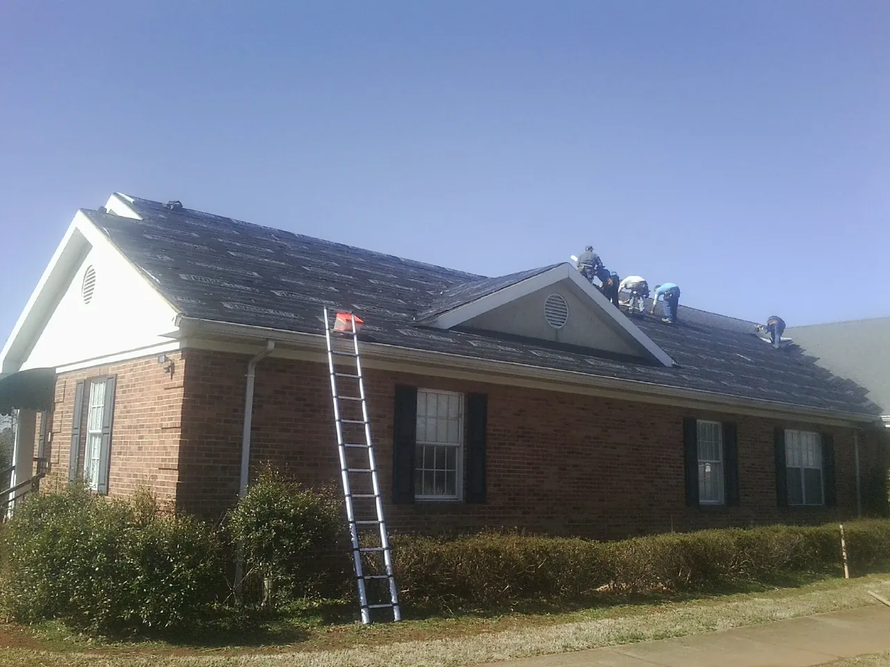 Crew tearing shingles off a brick ranch roof with a ladder set — Emergency Roof Repair work by our crew in King Hill