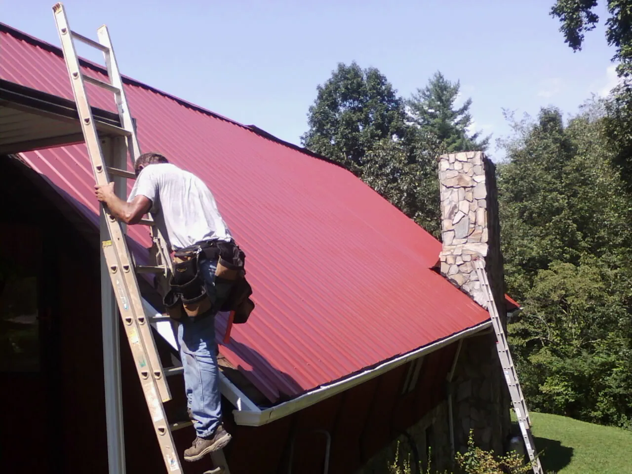 Roofer on a ladder beside a red metal roof and stone chimney — Soffit & Fascia Repair by Roofing Brothers in King Hill